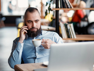 Young serious bearded businessman working on computer at table,drinking coffee.Man analyzes information, data, develops business plan. Freelancer, entrepreneur.Online marketing, education, e-learning