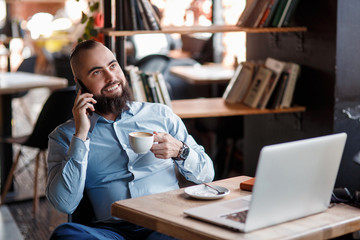 Young smiling bearded businessman is sitting at table in front of computer, talking on cell phone, holding pen. Freelancer works at home. Telephone conversations. Distance work, online education.