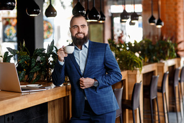 Young serious bearded businessman working on computer at table,drinking coffee.Man analyzes information, data, develops business plan. Freelancer, entrepreneur.Online marketing, education, e-learning