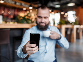 In the morning, the businessman follows news, the stock price via the telephone and drinks coffee. young investor. Bearded man with a smile looks into the phone with coffee in hand