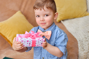Cute little boy gentleman in a blue denim shirt and peach bow tie holds a beautiful gift box in hearts and with a pink bow.	
