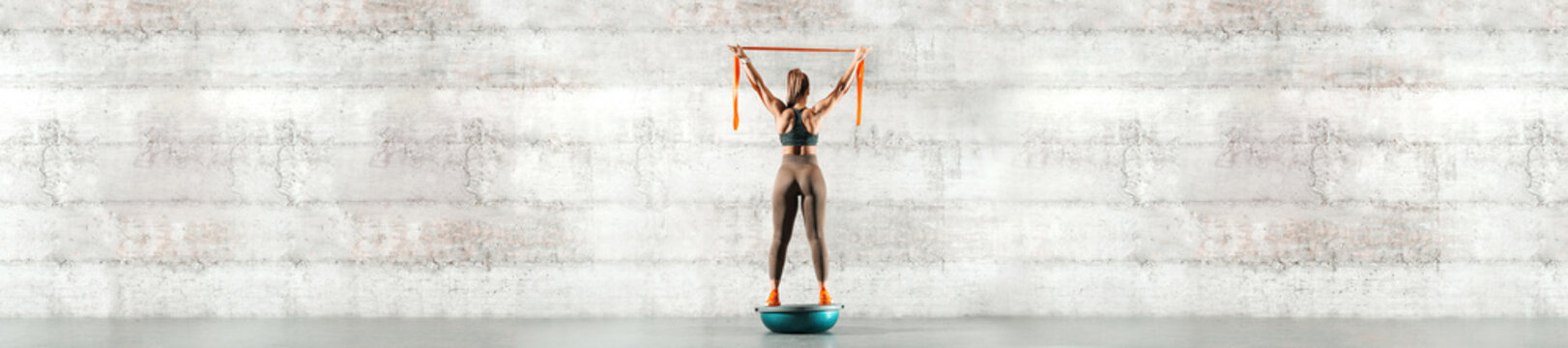 Full length of sporty woman standing on bosu ball and holding ribbon. Back turned, in background gray wall, copy space.