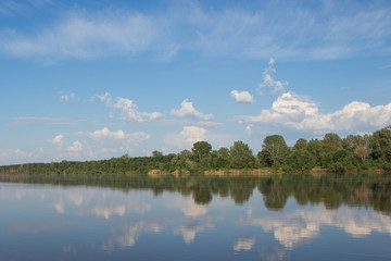 Summer. Blue sky, white clouds and green bank are reflected in the river. Symmetry.