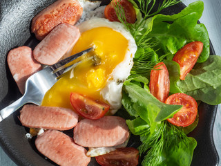 Morning sun illuminates the breakfast of fried eggs with sausages, cherry tomatoes and leaf lettuce on a black round pan, breakfast has already begun to eat. Serve on a wooden tray with a table fork.
