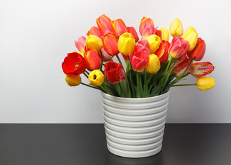 Huge bouquet of yellow and red tulips standing in a white large vase on a dark table against the background of a white wall