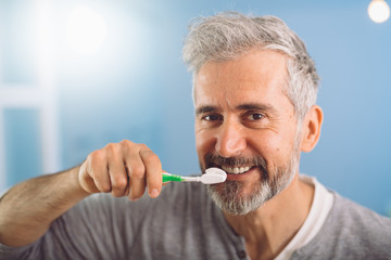 closeup of middle aged bearded gray haired man brushing his teeth against blue background