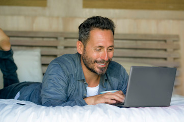 interior portrait of young attractive and happy man at home working relaxed on bed with laptop computer smiling cheerful in freelance networking business success