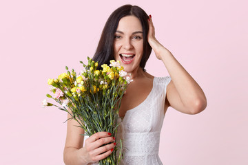 Half length portrait of pretty brunette woman holding bunch of flowers, laughing while looking directly at camera, keeps hand on head, dressed white dress, isolated over rosy studio background.