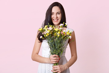 Image of girl holding bouquet of flowers in the hands, chorming lady expresses happyness, looking directly at camera, being in good mood on her birthday. Copy space for promotion or advertisment.