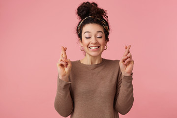 Portrait of a young girl with dark curly hair, crossed her fingers and closed eyes, hope for good luck. Isolated over pink background.