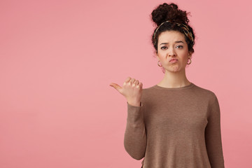 Frowning upset young beautiful girl with curly dark hair, pointing with finger to copy space...