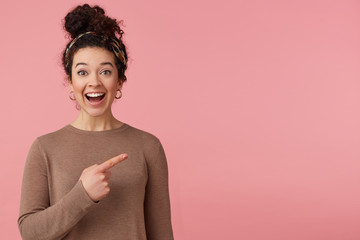 Portrait of happy amazed young beautiful lady with curly dark hair, heard the cool news, broadly smiling, looking at the camera, pointing with finger to copy space isolated over pink background.