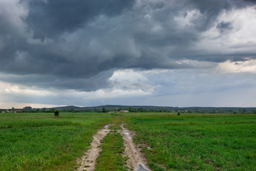 Fototapeta na wymiar Dirt road through green fields and storm clouds on the sky