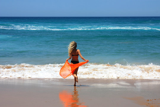 blonde in black bikini with orange pareo in her hands runs along the sand in an oncoming wave on a beach in northern Spain