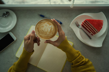Cup of cappuccino in woman hands with cake in cafe. Hot drink. Cozy coffee shop atmosphere in autumn or winter. Autumn or winter relax concept. Warm vintage toning. Selective focus.