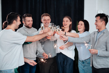 A group of young people celebrate the birthday of one of their friends. A young asian man is blowing candles on the cake. Sparkles in hands happy friends