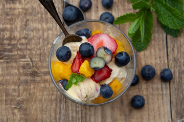 Fruit salad with ice cream - blueberries, strawberries, mango and mint, summer refreshing dessert. Wooden background. Glass with vanilla icecream.