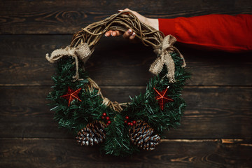 Christmas holiday background. Celebration theme. Celebration concept, free space for text. Female hand with christmas wreath with fir, pine cones, red berry and ivy on brown rustic background.