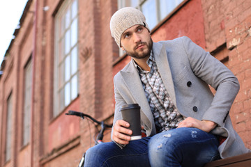 Fototapeta na wymiar Handsome young man in grey coat and hat sitting on a bench relaxed drinking coffee and thinking near his bicycle.