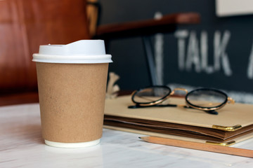 closeup paper cup of hot coffee, notebook and glasses on white table in cafe with soft-focus and over light in the background