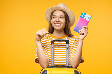 Close-up studio portrait of dreaming young female tourist holding flight tickets and suitcase,...