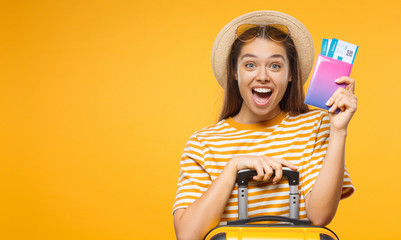 Horizontal banner of happy surprised young female tourist  holding suitcase passport with flight tickets, isolated on yellow background with copy space - obrazy, fototapety, plakaty