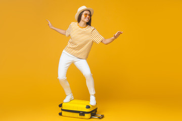 Fototapeta na wymiar Young tourist girl standing on suitcase, pretending like she is surfing, isolated on yellow background. Dream about traveling concept