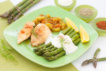 Roasted chicken breast meat and grilled asparagus with sauce on green plate. Asparagus meal decorated with lemon and dill.