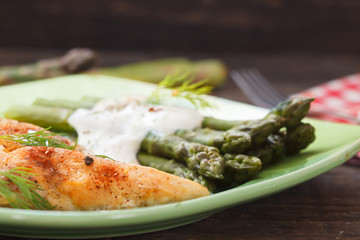 Roasted chicken breast meat and asparagus with sauce on green plate. Asparagus meal decorated with lemon and dill. Homemade cuisine. Close up image, wooden background.