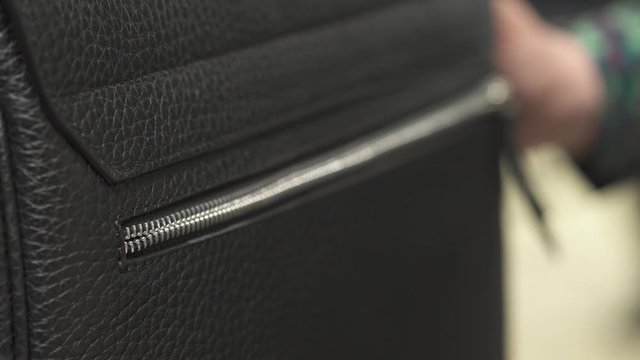 A close-up demonstration video of woman's hand with nude manicure opening zipper of male black leather bag. Isolated, on blurred background