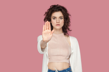 Stop. Portrait of serious beautiful brunette young woman with curly hairstyle in casual style standing with stop hands gesture and caution to dont touch. indoor studio shot isolated on pink background