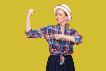 Strong and independent alone. Portrait of proud modern stylish mature woman in casual style with white hat standing and pointing finger at her biceps. indoor studio shot isolated on yellow background.