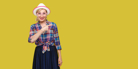 Portrait of happy modern stylish mature woman in casual style with hat and eyeglasses standing looking at camera and pointing and wall empty copyspace. indoor studio shot isolated on yellow background