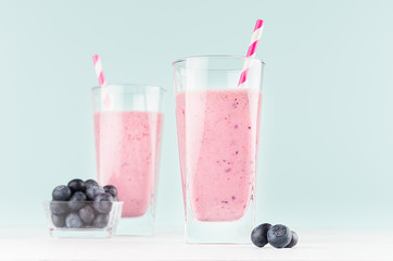 Pink smoothies with blueberries in bowl, striped straws in glass closeup on white wood table and pastel blue wall, copy space.