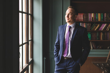 Fototapeta premium Portrait of a successful businessman standing in an expensive business suit near the window in his office and smiling