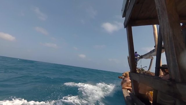 Traditional wooden Dhow boat with tourists sailing fast through ocean waves on bright sunny day. Sea horizon with tropical island and blue sky in the background. View from the left side of the boat.