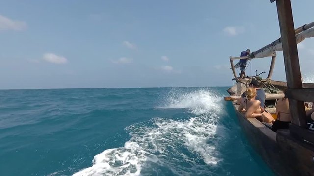 Traditional wooden Dhow boat with tourists sailing fast through ocean waves on bright sunny day. Sea horizon with tropical island and blue sky in the background. View from the left side of the boat.