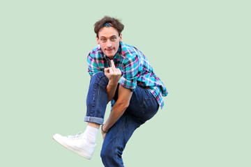 Portrait of angry young man in casual blue checkered shirt and headband standing, looking at camera and showing middle finger fuck sign. indoor studio shot, isolated on light green background.