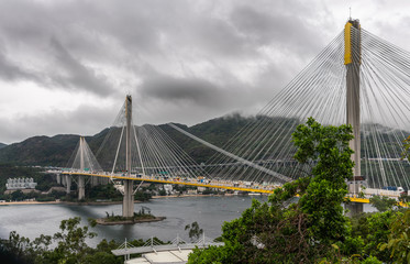 Hong Kong, China - March 7, 2019: Morning under dark rainy sky. Ting Kau Bridge linking New Territories from Wok Tai Wan, Kowloon. High rise buildings, dark hills on horizon, Green foliage up front.