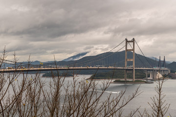 Hong Kong, China - March 7, 2019: Morning under dark rainy sky. Tsing Ma Bridge crossing gray Ma Wan Channel from Wok Tai Wan to Park Island. Dark hills on horizon.