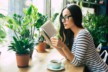 Happy beautiful young woman in glasses is reading book and drinking coffee in modern cafe