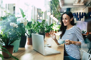 Beautiful happy young woman using laptop and drinking cup of coffee in cafe while lunch time