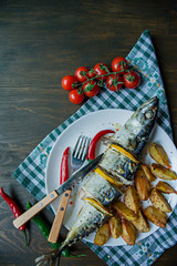 Baked mackerel with lemon and baked potatoes on a white plate. Fresh vegetables . Cherry, chili pepper. Cutlery. View from above. Wooden table. Space for text.