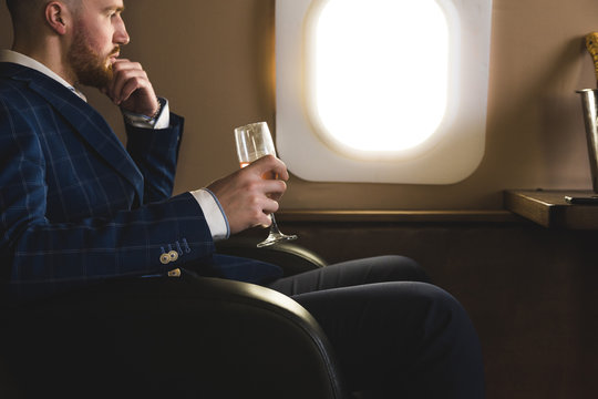 A young successful businessman in an expensive suit sits in the chair of a private jet with a glass of champagne in his hand and looks out the window