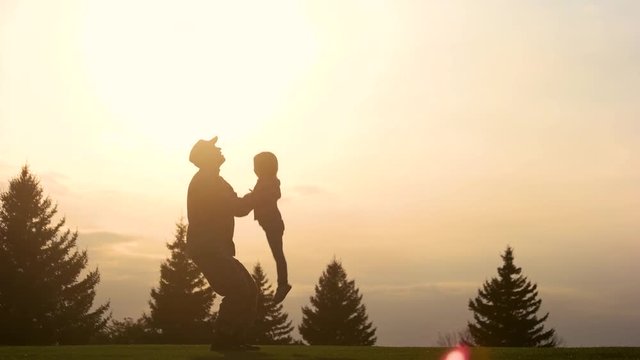 Playing with daughter in the evening park. Silhouette of military father lifting up and down little girl into the air.