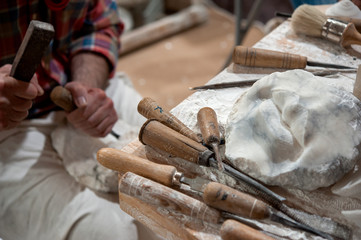  Sculptor working on alabaster sculpture in his workshop with hammer and chisel. Various tools on the bench.