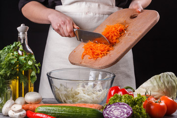 young woman in a gray apron adds carrot to a salad