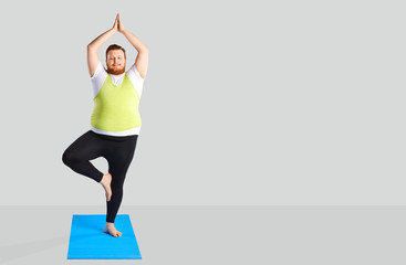 Funny fat man doing yoga exercises standing on a gray background.
