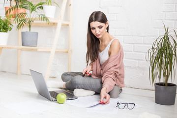Young woman sits on the floor in a Scandinavian apartment interior with a laptop, studying law,...