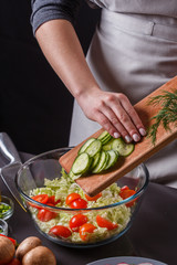 A young woman in a gray apron adds a cucumber to a salad